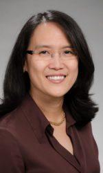 A photo of Dr. Janie Lee. Dr. Lee is wearing a brown blouse with a gold necklace and glasses. She smiles at the camera in front of a neutral grey background.