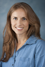 A headshot of Dr. Sarah Menashe. Dr. Menashe wears a blue button-down shirt and a big smile. She has long auburn hair.