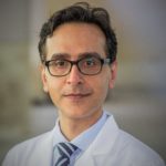 A photo of Dr. Amir Iravani. Dr. Iravani wears a lab coat, button up shirt and glasses while looking directly into the camera.