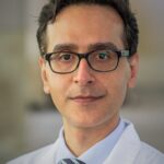 A photo of Dr. Amir Iravani. Dr. Iravani wears a lab coat, button up shirt and glasses while looking directly into the camera.
