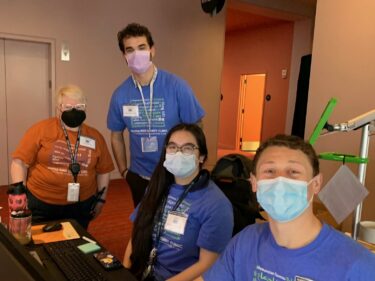 Medical professionals Le Ann, Lukas Wood, Janelle Dao and Brandon Morales pose for a group photo. They wear t-shirts and medical masks while gazing at the camera.