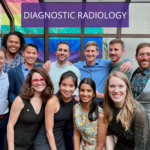 Diagnostic radiology graduates pose for a group picture in front of a rainbow-colored flag. They are dressed up, smiling and gazing into the camera.