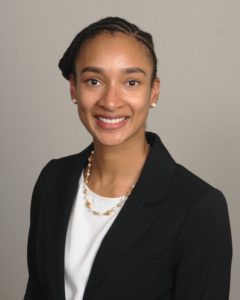 A photo of Marissa Lawson. Marissa wears a dark-colored blazer with a white shell and gold necklace. She smiles into the camera in front of a neutral grey background.