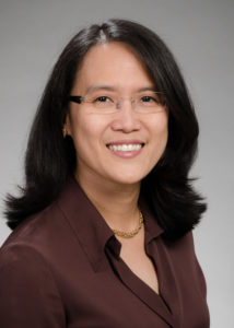 A photo of Dr. Janie Lee. Dr. Lee is wearing a brown blouse with a gold necklace and glasses. She smiles at the camera in front of a neutral grey background.