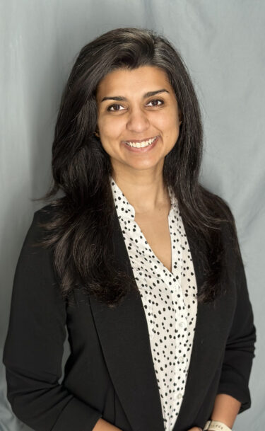 Dr. Anum Kazerouni wears a light colored blouse with a polka dot pattern on it and a black blazer. she has long dark hair and smiles at the camera.