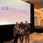 Doctors Partridge, Kazerouni and Rahbar pose smiling for the camera in front of a projection screen displaying ECOG-ACRIN logo