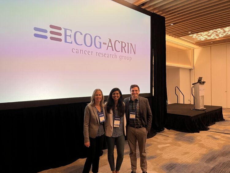 Drs Partridge, Kazerouni and Rahbar pose smiling for the camera in front of a projection screen displaying ECOG-ACRIN logo 