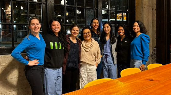 Members of the Women in Radiology group pose for a group photo. They where casual attire and stand in front of a window and behind a table.