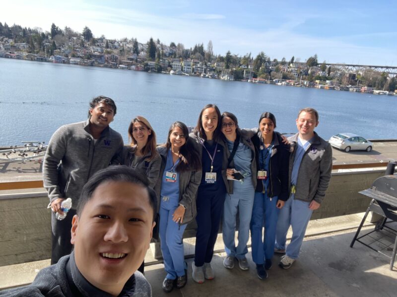 Trainees pose in front of the blue water of Portage Bay. They wear medical attire underneath jackets.