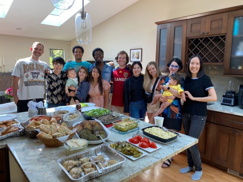 Trainees smile at the camera in a kitchenette behind a variety of potluck dishes.
