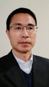 A headshot of Dr. Feng Zhang. In the photo, Dr. Zhang wears glasses and a button-up shirt underneath a sweater and blazer as well as a pleasant, serious expression.