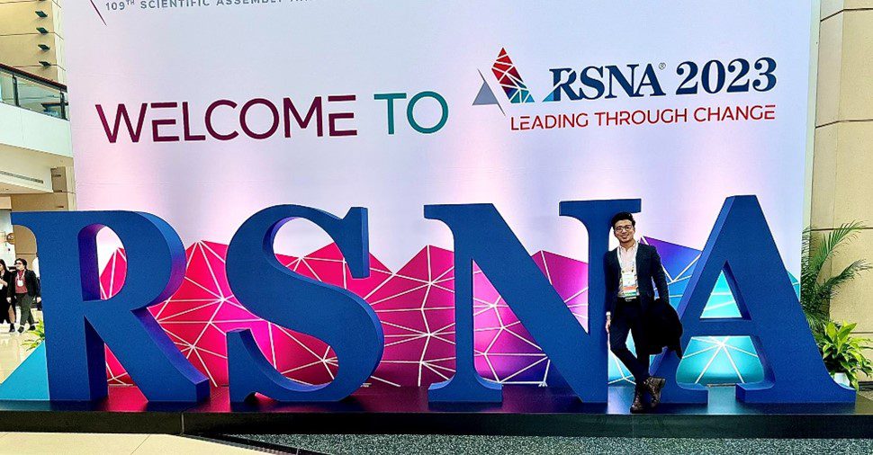 Saubhagya Srivastava, MBBS, poses for a photo in front of the RSNA sign wearing a black suit and looking relaxed.