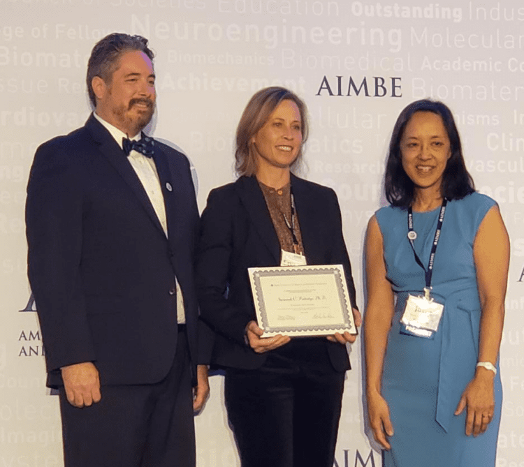 Dr. Savannah Partridge (center) holds a certificate while flanked by two other people. Dr. Partridge wears a black suit and lanyard and smiles broadly. The acronym AIMBE is printed on a sign behind her.
