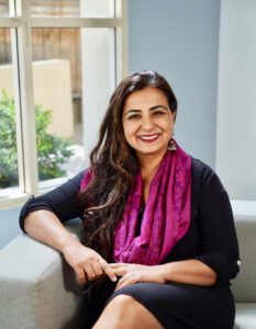 A photo of Dr. Bhavya Rehani. Dr. Rehani wears a black outfit with a bright pink scarf. She has long, dark hair, a big smile, and she holds her hands in a relaxed position in front a pastel-blue window frame.