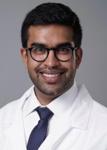 Dr. Nitin Venugopal wears a white button up, dark blue tie, and a white lab coat while smiling in front of a grey background.