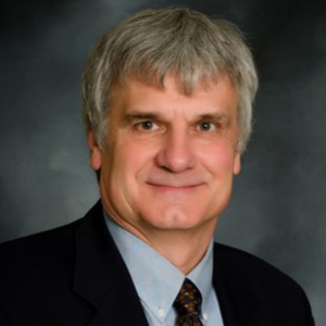 Dr. Bob Mach wears a light blue shirt and a black jacket, while smiling in front of a grey background.