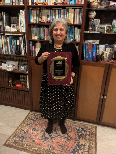 Dr. Kalpana Kanal poses with her award in front of a bookshelf. She wears a black outfit and smiles at the camera.